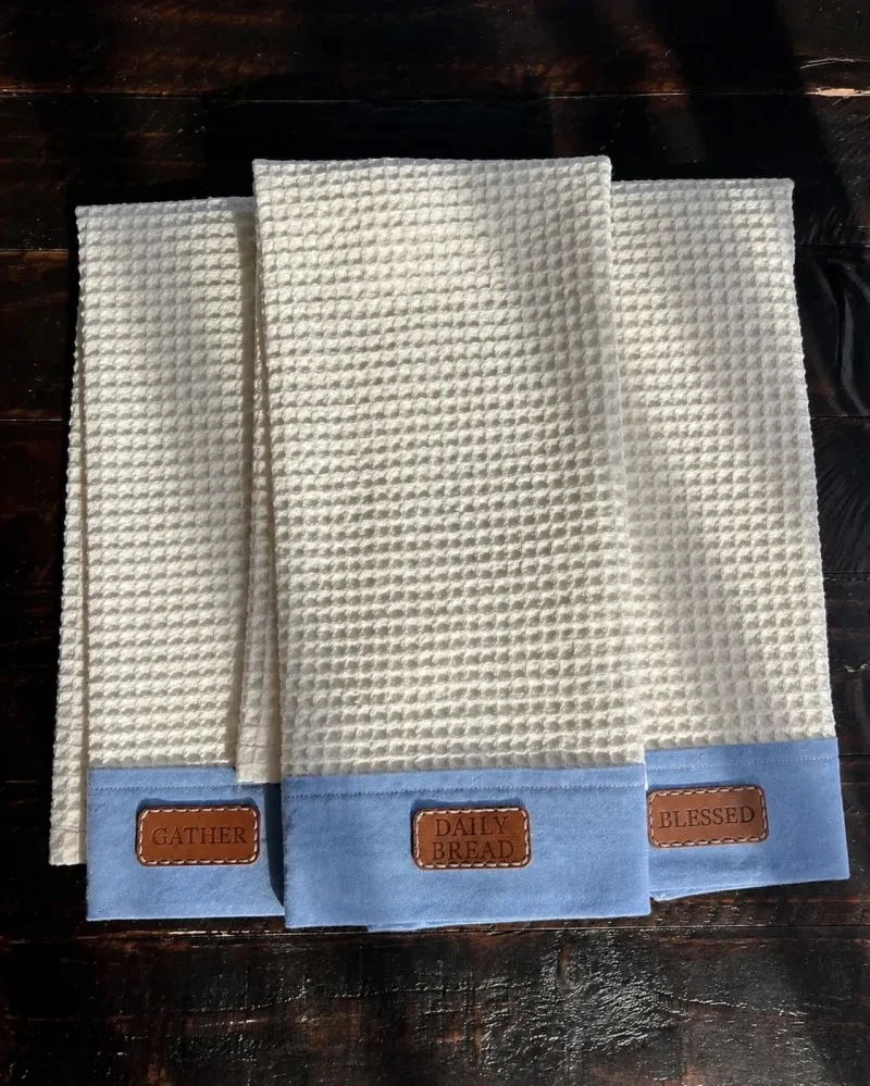 Three cream waffle-textured towels with blue chambray borders and Gather, Daily Bread, and Blessed patches